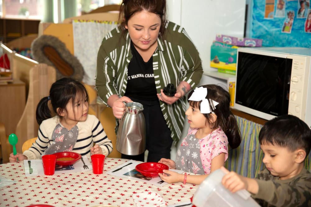Image of an adult serving children tea at a table in a playroom.