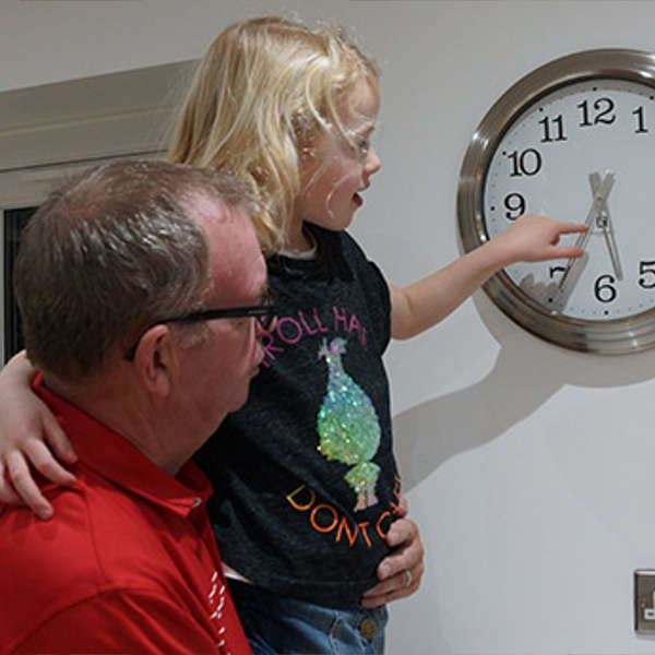 Image of an adult holding a child who is pointing at an analogue clock on the wall.