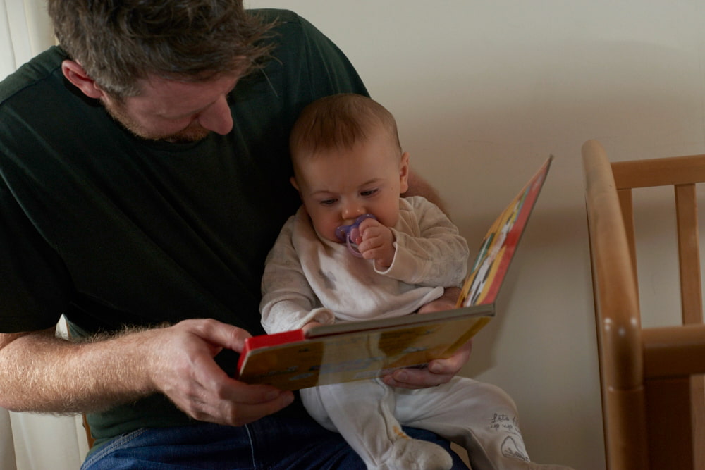 Image of a dad reading from a book with his baby sitting on his lap looking at the book.